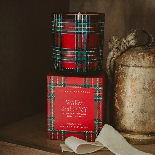 A red scented candle in a dark glass jar with a green and red tartan plaid pattern, displayed on top of its matching red and plaid box. The box is labeled 'WARM and COZY' by 'Sweet Water Decor'.