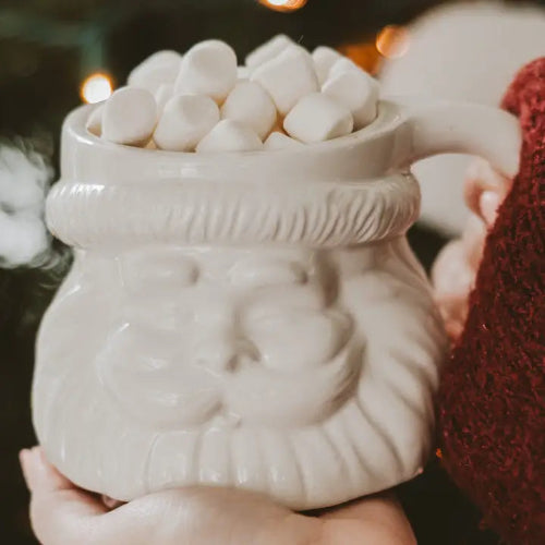 A Santa Claus mug filled with mini marshmallows, held in hands with a cozy, warm background.