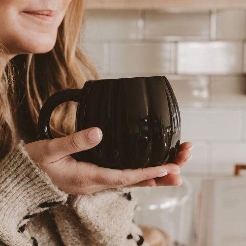A person in a beige knit sweater holds a glossy black ceramic mug in a warm, softly lit kitchen setting with white subway tiles in the background.