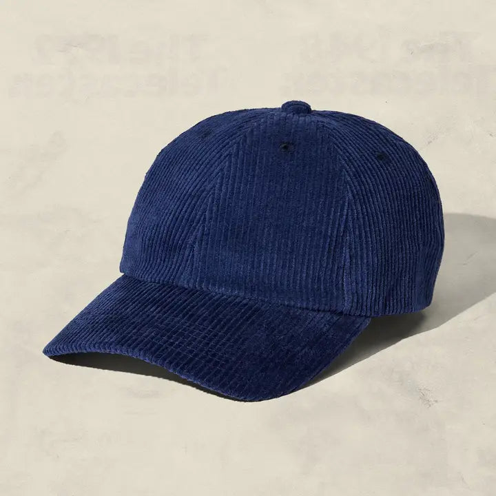 A dark blue corduroy baseball cap with a curved brim, photographed on a textured light beige background.