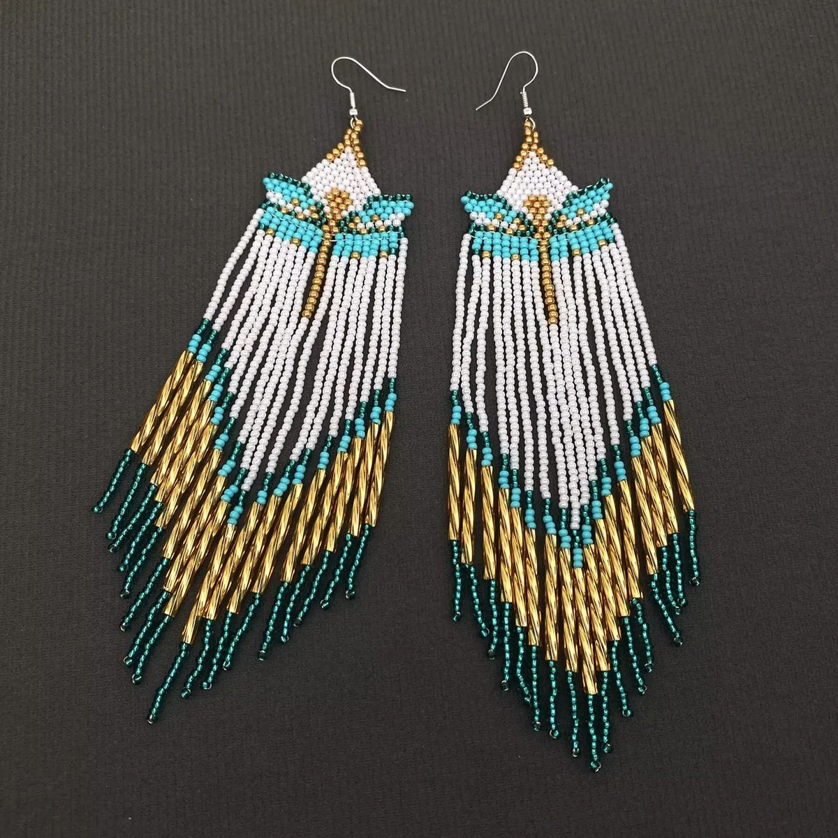 A pair of long beaded dangle earrings with white, turquoise, and gold seed beads, hanging against a dark textured background.