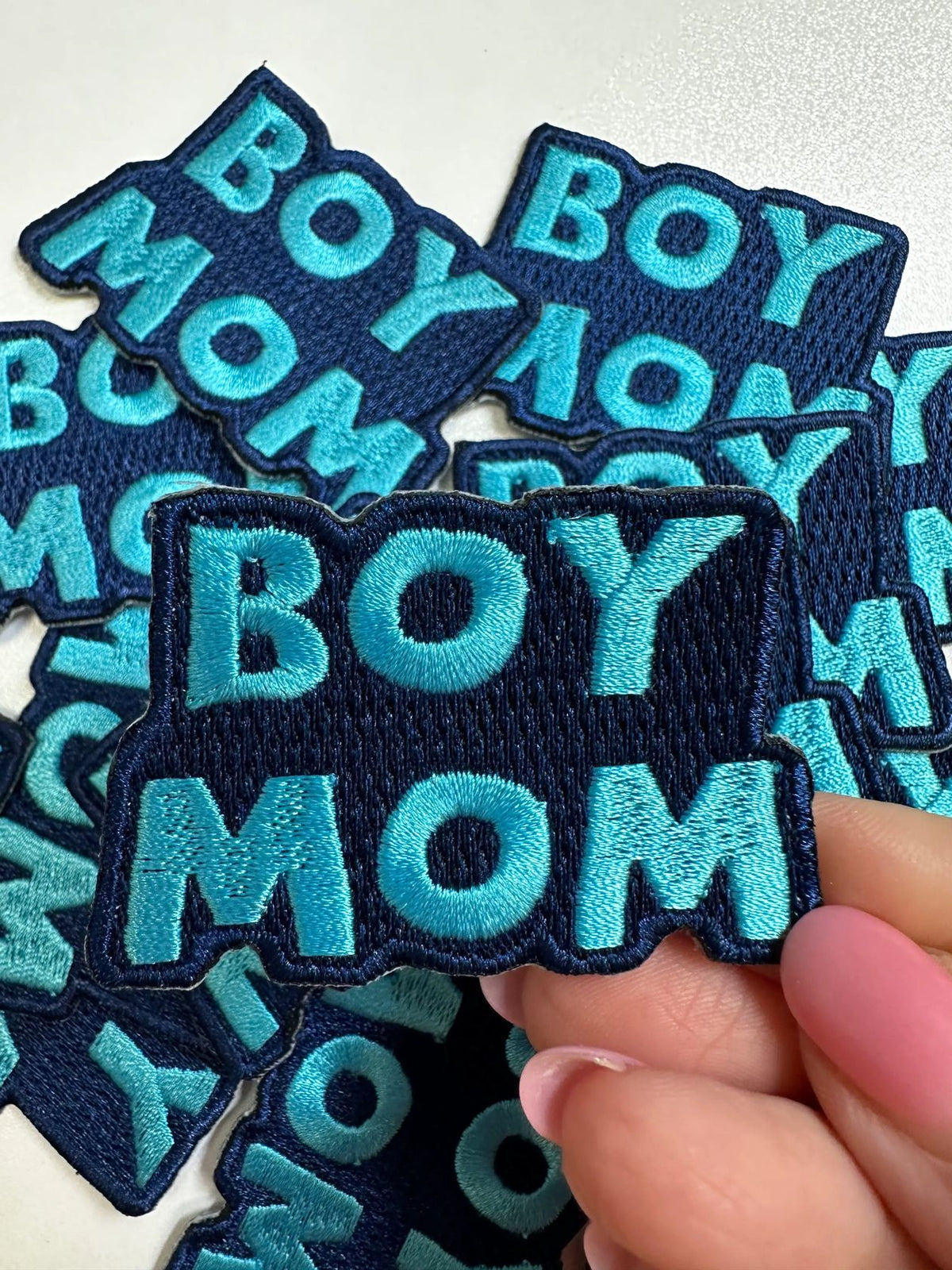 A hand holding an embroidered patch that reads 'BOY MOM' in bright turquoise lettering on a dark navy background, with several matching patches scattered behind it.
