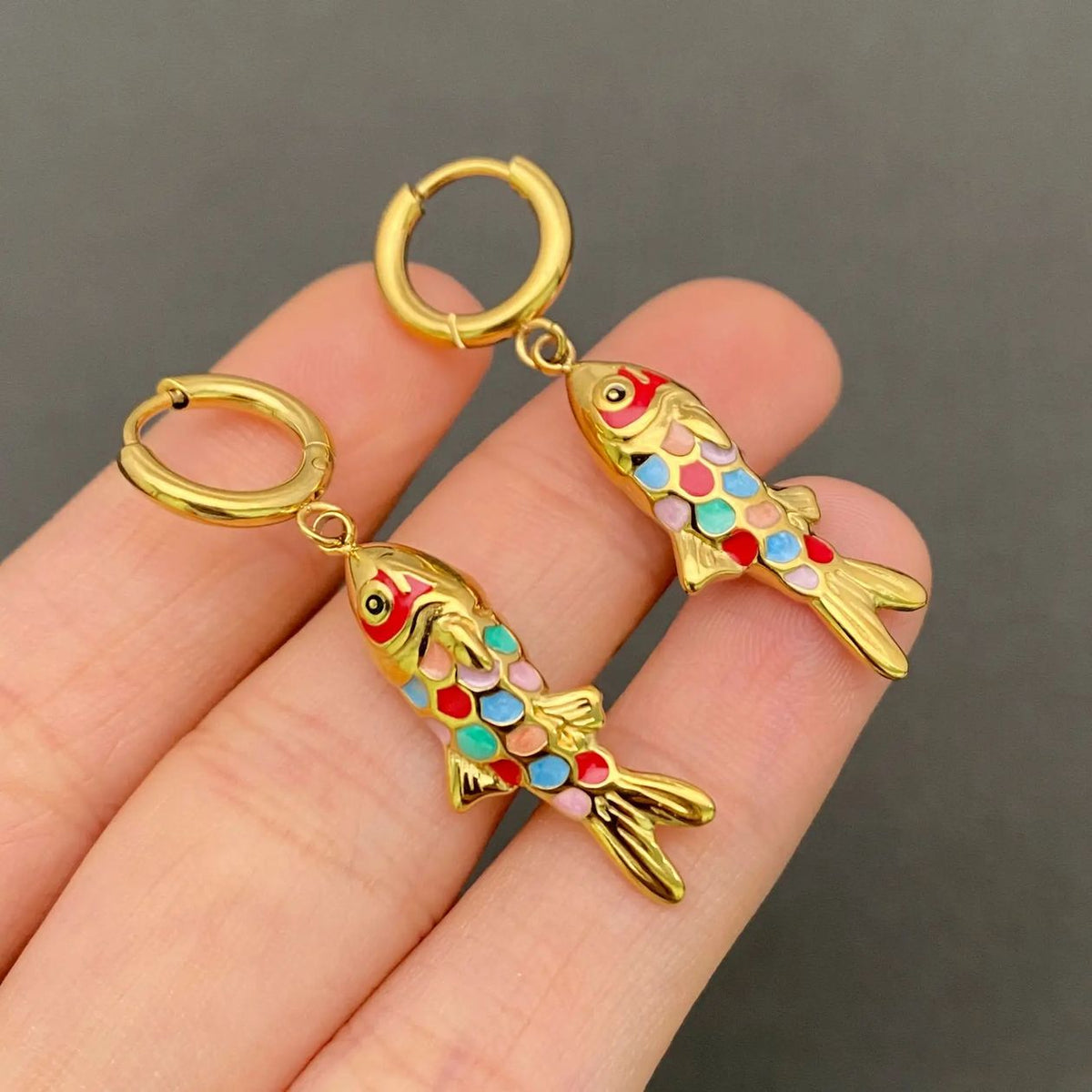 A pair of gold fish-shaped huggie hoop earrings with colorful enamel details, held on a person's fingers against a dark gray background.