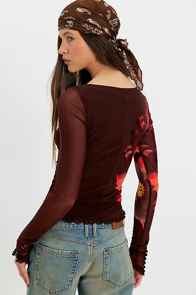 A woman shown from the back wearing a sheer long-sleeve top in dark brown with a red and orange floral print on one sleeve, paired with blue jeans and a brown patterned bandana.