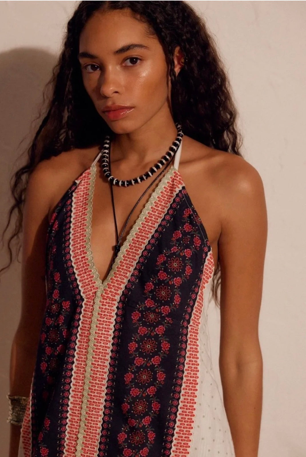 A woman wearing a sleeveless halter-style dress with a dark navy base, red and beige geometric border patterns, and a red floral print, styled with layered necklaces against a plain beige wall.