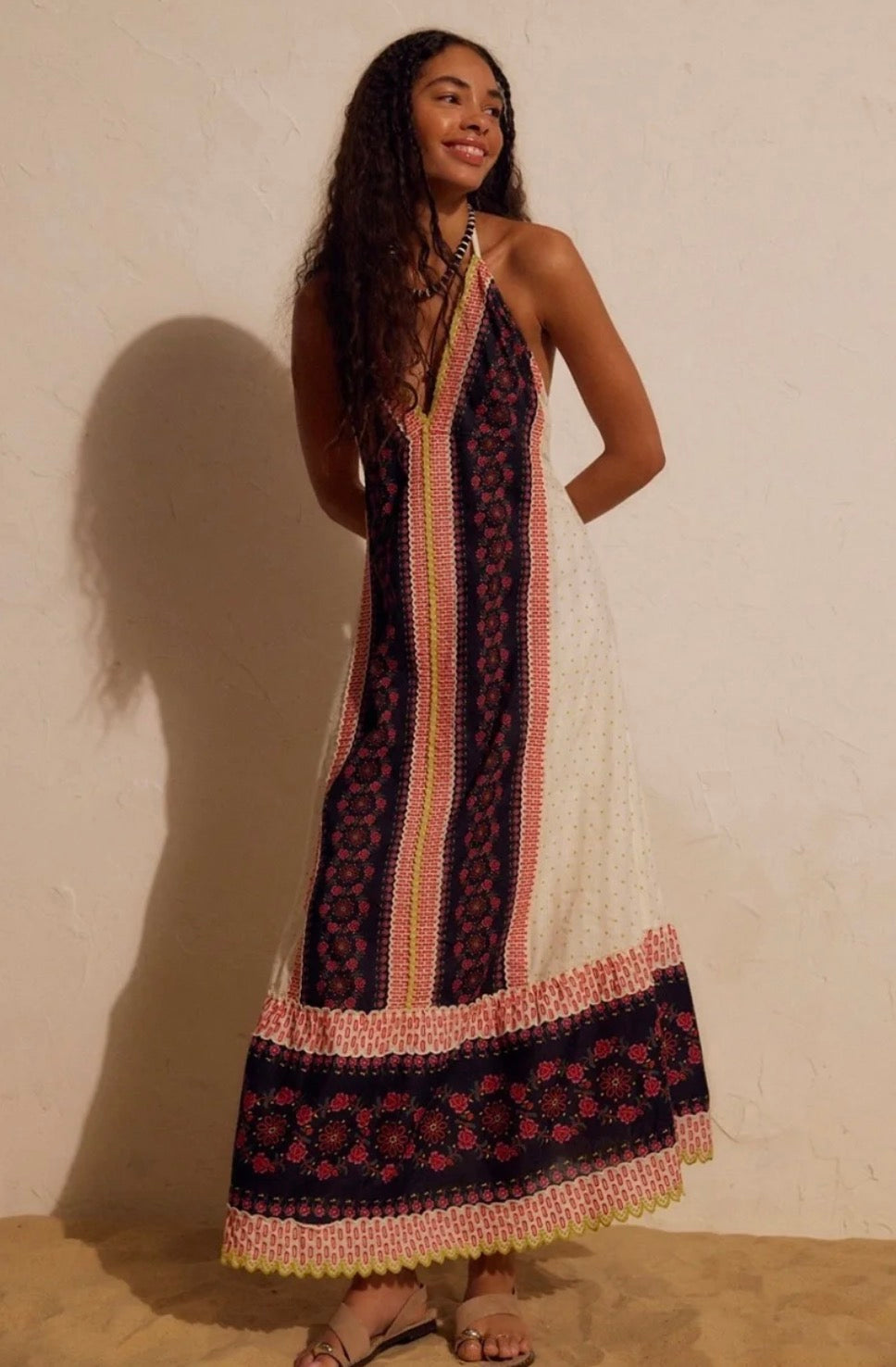 A woman modeling a sleeveless maxi dress with a bohemian patterned design in navy, pink, and cream, standing against a textured beige wall.