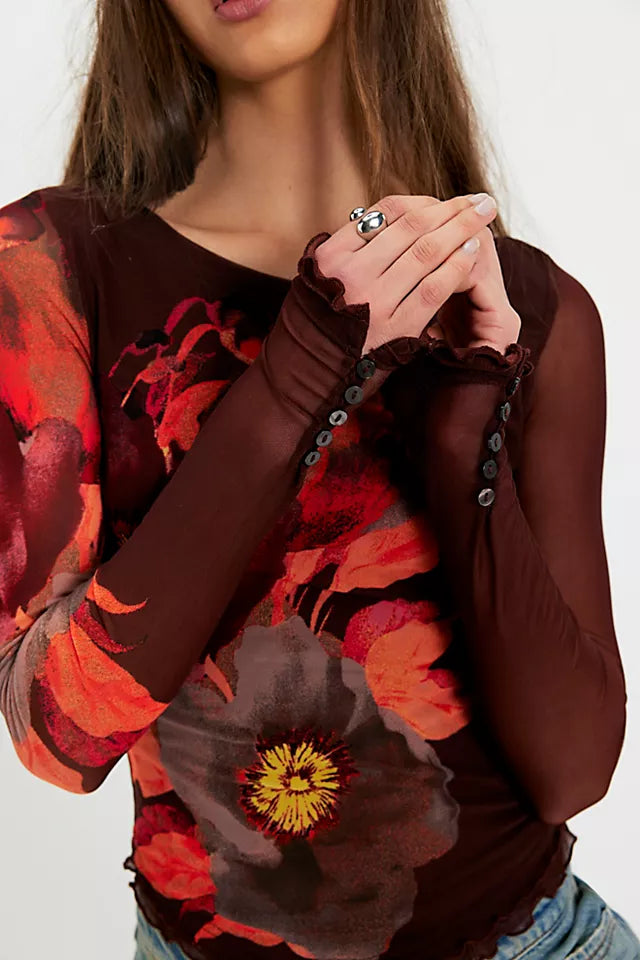 A close-up of a woman wearing a sheer long-sleeve top in dark brown with a large red and orange floral print and ruffled cuffs.