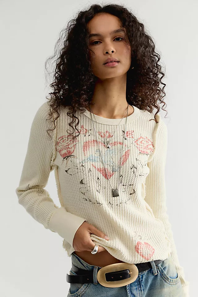 A woman with curly dark hair wearing a cream-colored waffle-knit sweater with a pink and black graphic print, paired with blue jeans and a black belt with a gold buckle, against a plain light gray background.