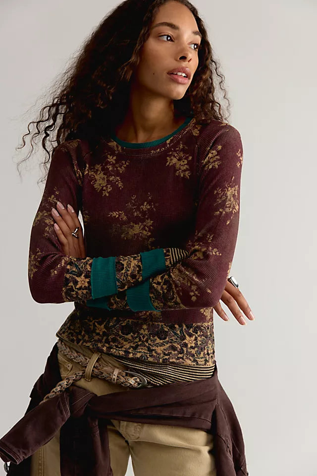 A woman modeling a long-sleeve crewneck top in deep burgundy with a gold floral pattern, teal ribbed cuffs, and a matching patterned waist sash, paired with khaki pants against a light gray studio background.