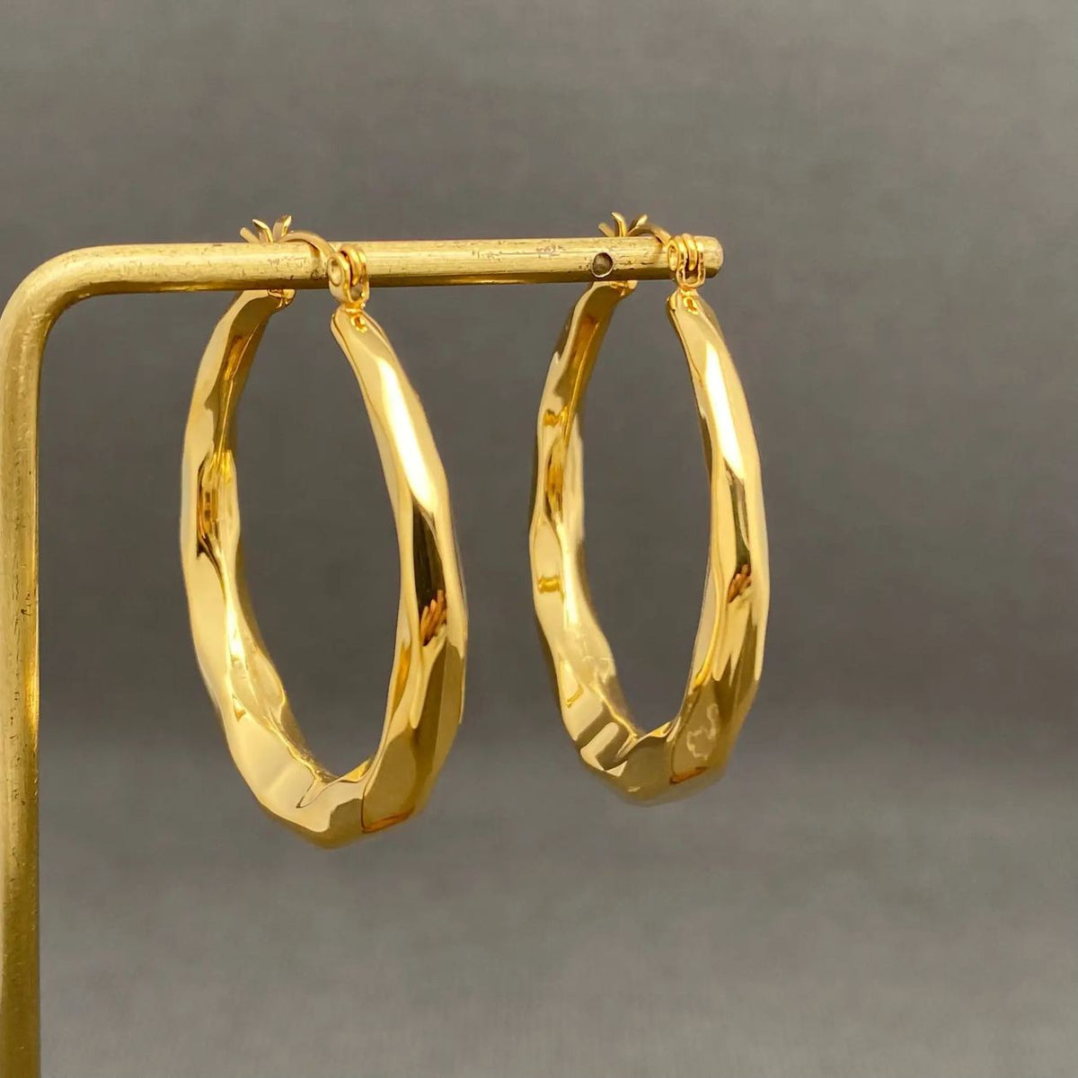 Pair of large, polished gold-plated geometric hoop earrings displayed on a gold-colored metal stand against a neutral gray background