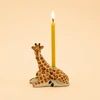 A novelty candle shaped like a giraffe with a lit yellow candle inserted, shown against a plain beige background.