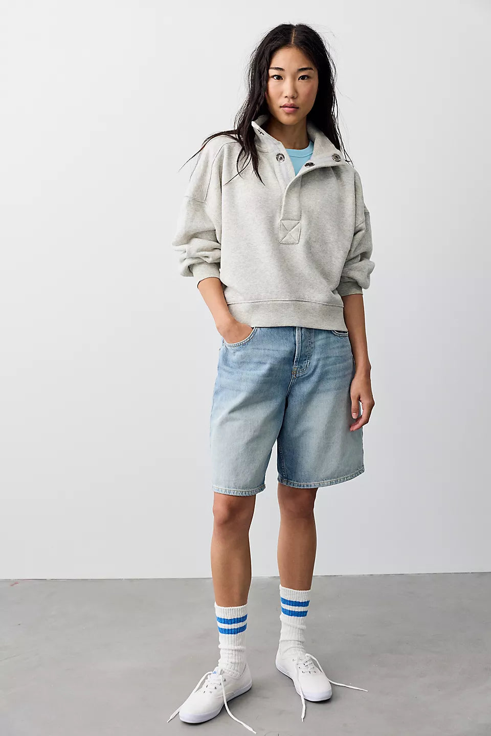 A model stands in a studio wearing a light gray oversized hoodie, light-wash denim shorts, white crew socks with blue stripes, and white sneakers against a plain light gray background.