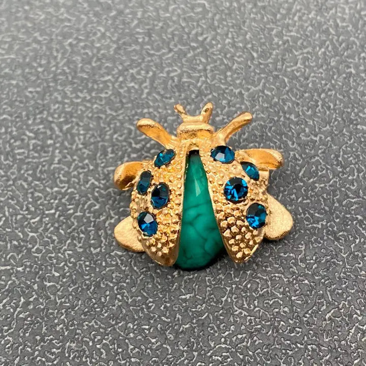 A gold ladybug-shaped brooch or charm with a polished green body and blue rhinestone accents, photographed on a textured gray background.
