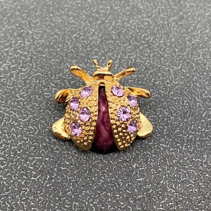 A gold ladybug-shaped brooch or charm with purple rhinestones and a dark purple center, shown on a textured gray background.