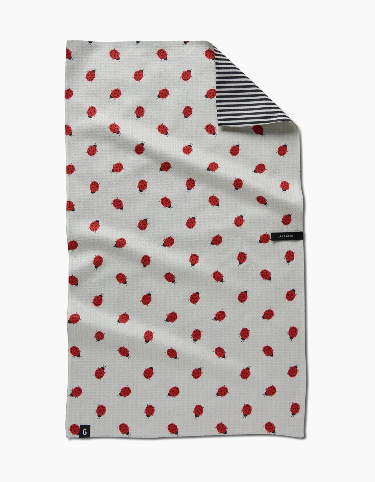 A white waffle-weave kitchen towel with a red ladybug pattern, featuring a striped black and white reverse side and small black brand labels.