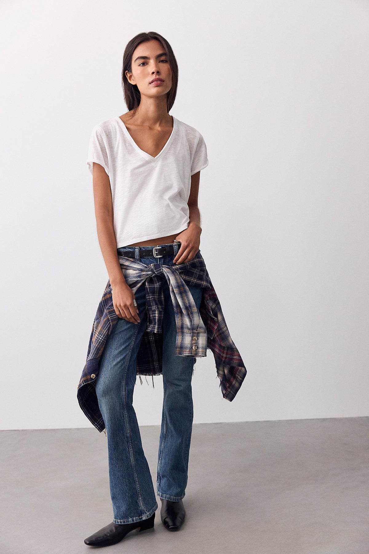 Model wearing We The Free Monte Low-Rise Slim Bootcut Jeans in Twilight with a white tee and plaid shirt tied at the waist