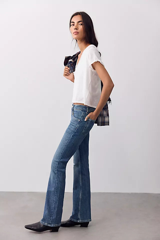 A woman modeling blue flare jeans with a white t-shirt and black boots, holding a plaid shirt under her arm against a plain studio background.