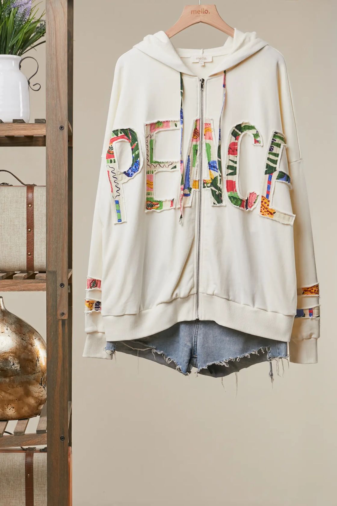 An oversized cream zip-up hoodie with colorful patchwork 'PEACE' lettering across the front, hanging on a wooden hanger against a beige wall. The hoodie features multicolor patterned details on the sleeves and under the hem, styled with frayed denim shorts.