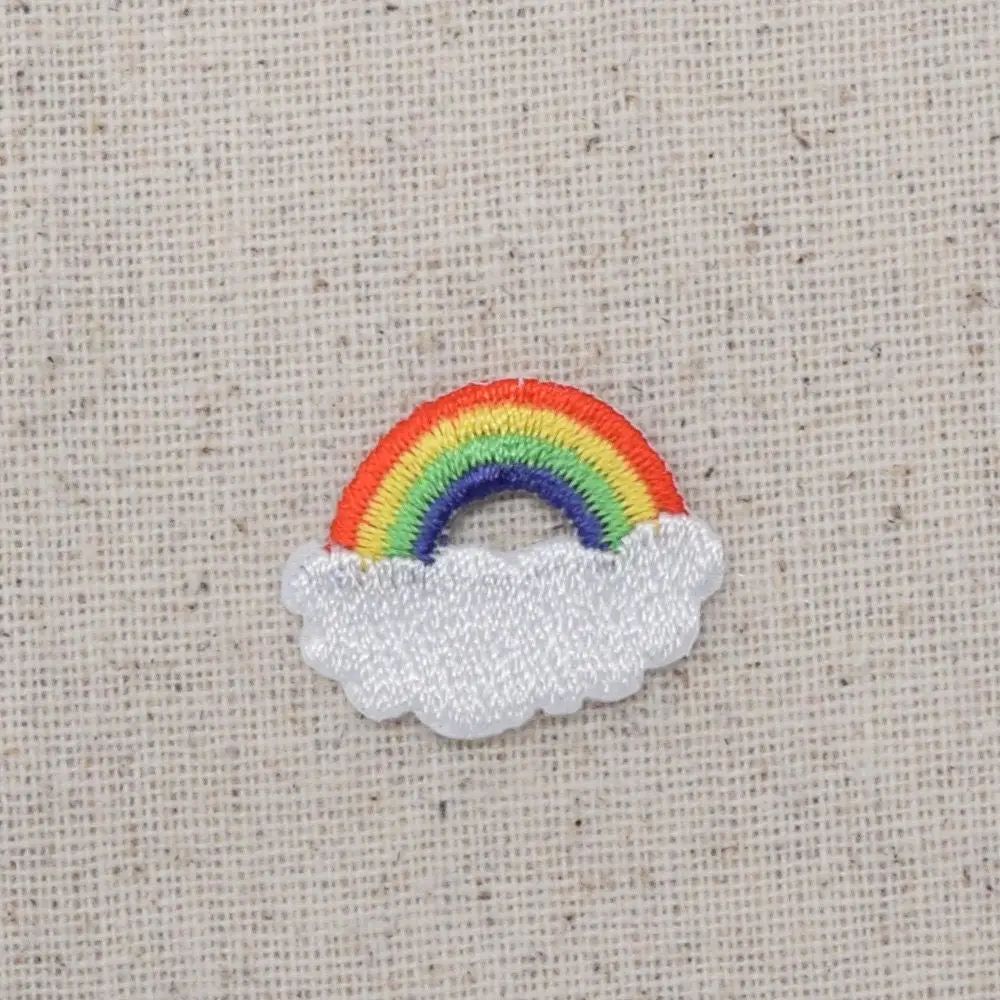 An embroidered patch featuring a small rainbow arching over a fluffy white cloud, displayed on a textured beige linen background.