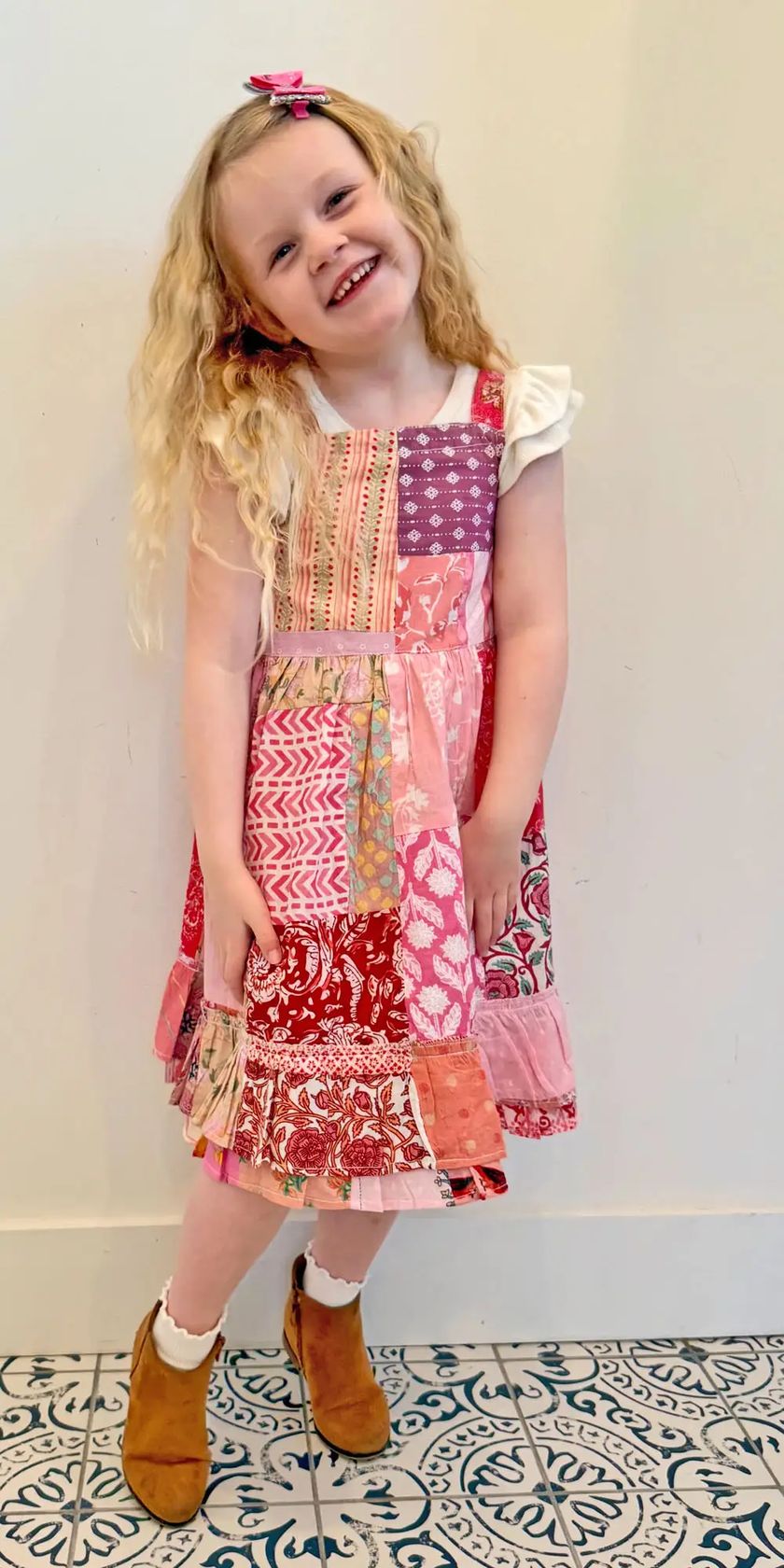 A young girl with long blonde hair wearing a colorful patchwork-style dress with pink, red, and purple patterns, white ruffled sleeves, brown boots, and white socks, standing on a patterned tile floor against a plain wall.