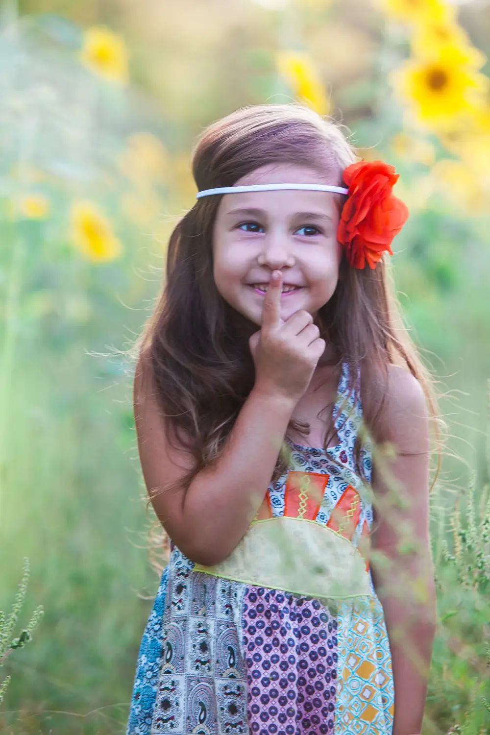 A young girl with long brown hair wearing a colorful patchwork dress and a red flower headband, posing in a sunflower field.
