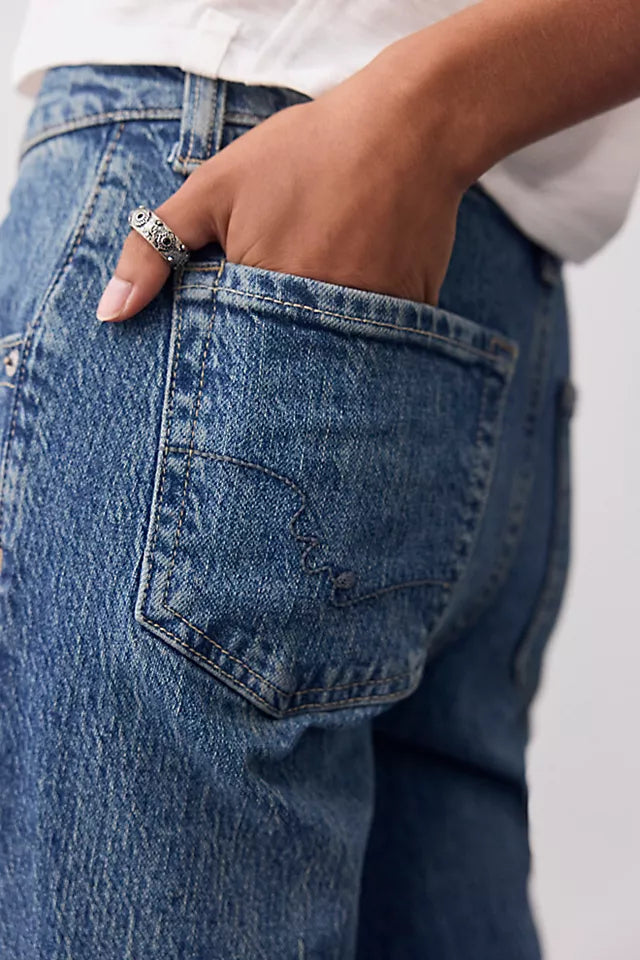 Close-up of a person wearing blue denim jeans, with the back pocket and hand in the pocket visible. A silver ring is worn on the hand, and a white shirt is partially visible at the top.