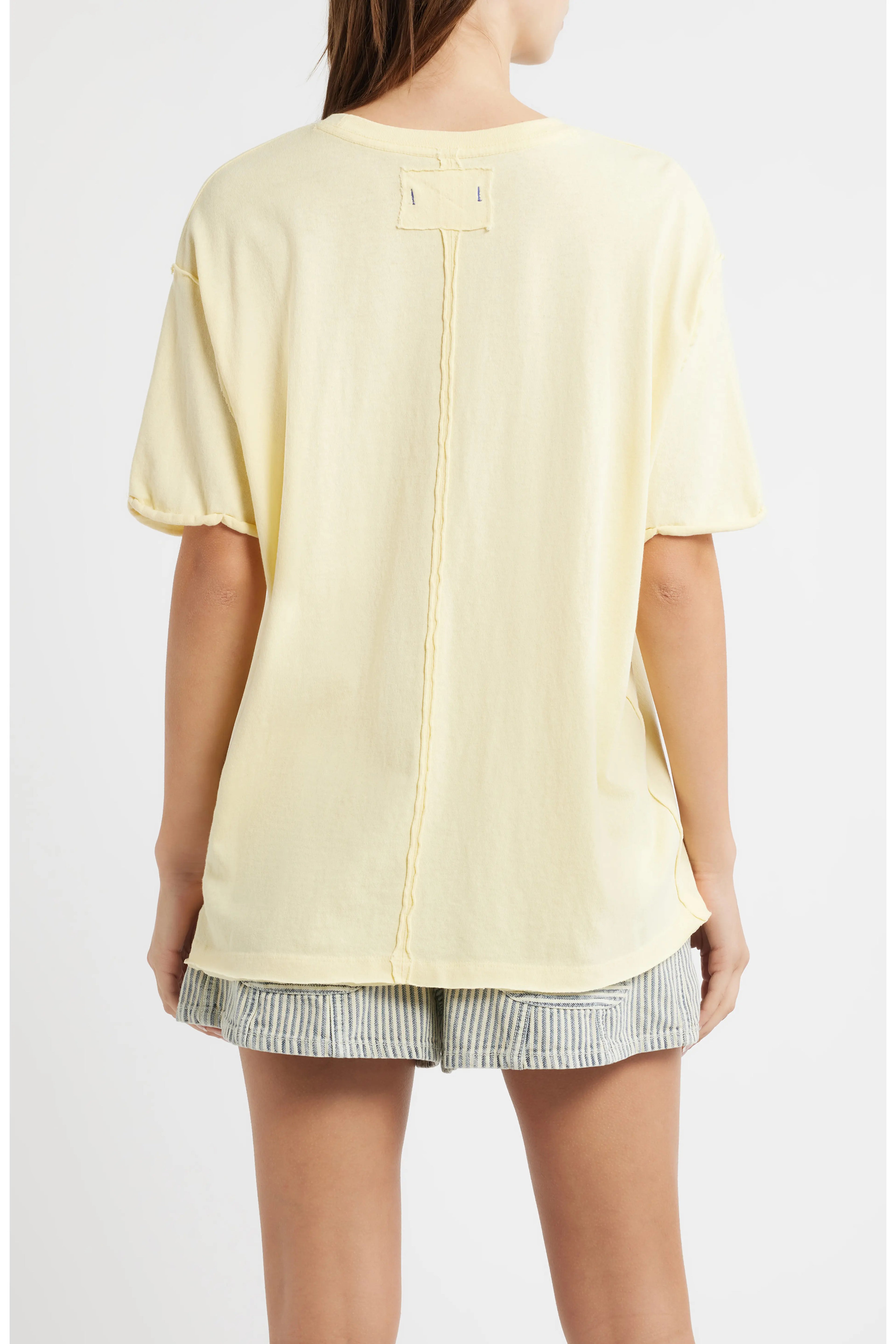 A woman shown from the back wearing a pale yellow oversized short-sleeve top with a vertical seam detail and striped shorts against a light gray studio background.
