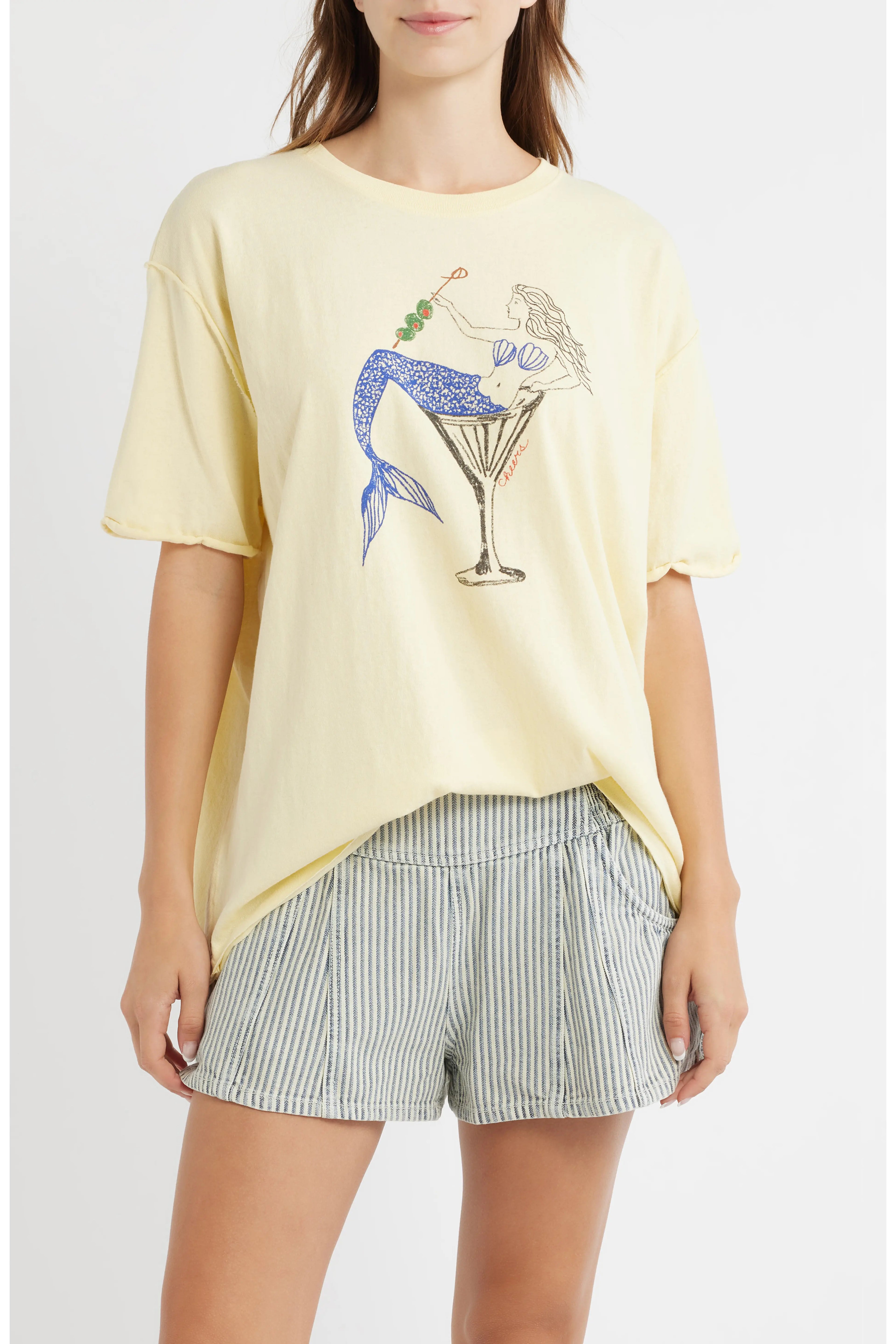 A woman wearing a pale yellow graphic t-shirt with a mermaid in a martini glass design, paired with blue and white striped shorts, against a light gray studio background.