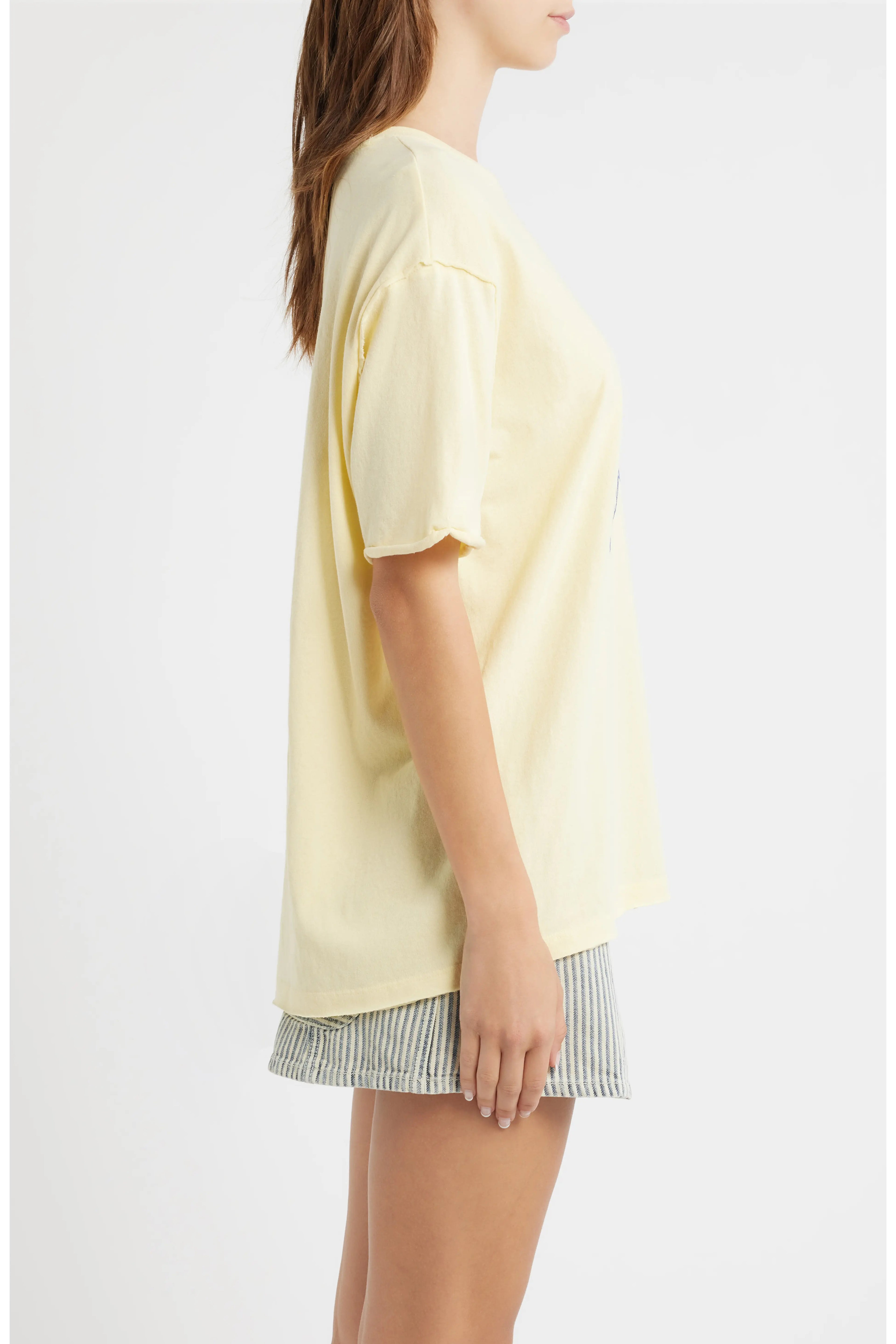A woman shown in side profile wearing an oversized pale yellow short-sleeve T-shirt and striped shorts against a plain light gray studio background.