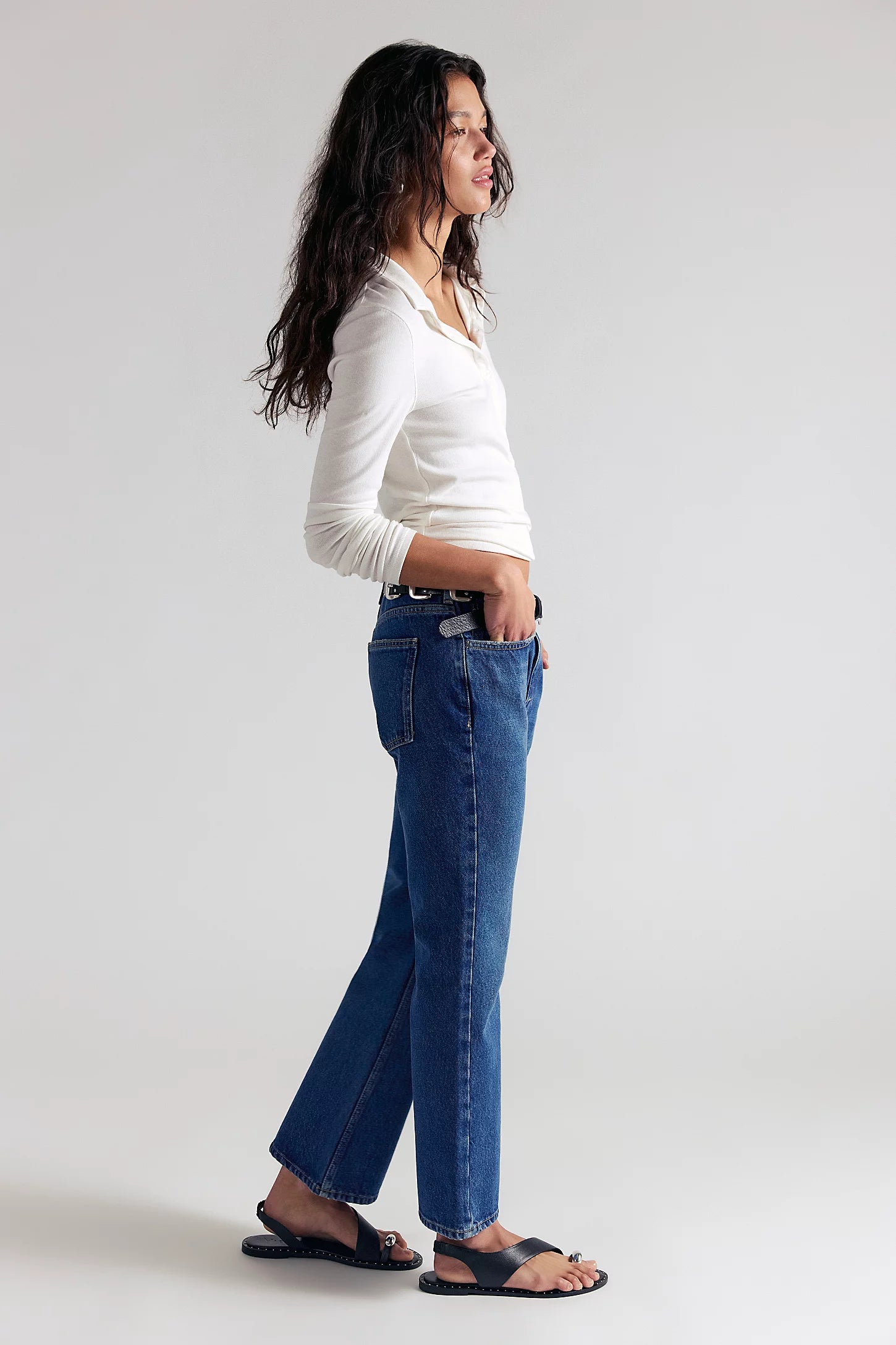 A woman in a white long-sleeve top and blue jeans stands in profile against a light gray studio background, wearing black sandals and a black belt.