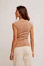 A woman shown from the back wearing a fitted sleeveless top in a warm beige or tan color, paired with light cream pants, against a plain light gray studio background.