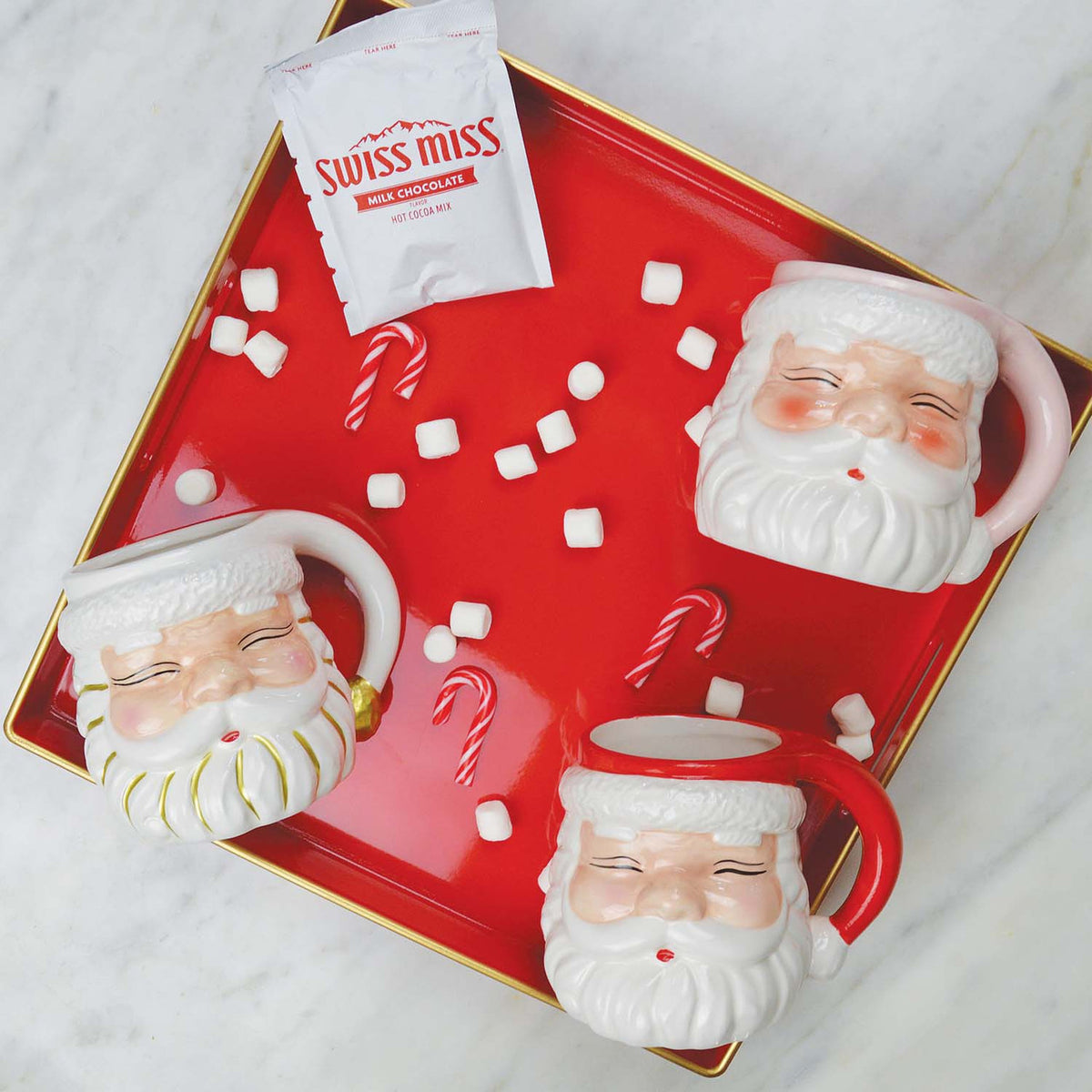 White and red ceramic Santa mugs on a red tray with mini marshmallows, candy canes, and hot cocoa