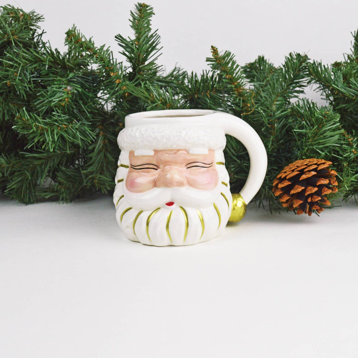 White ceramic Santa coffee mug with rosy cheeks styled with evergreen garland and a pinecone