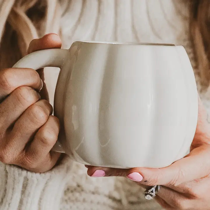 A person holding a large, cream-colored ceramic mug with a ribbed pumpkin-like design. The person is wearing a cozy white knit sweater, and a silver ring is visible on one of their fingers.