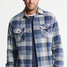 A man wearing a blue and white plaid button-up flannel shirt over a navy crewneck t-shirt, shown from the chin down against a plain white background.