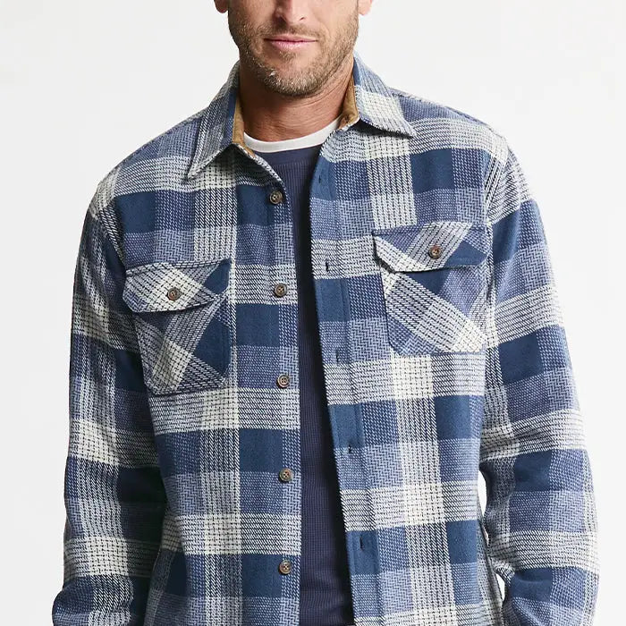 A man wearing a blue and white plaid button-up flannel shirt over a navy crewneck t-shirt, shown from the chin down against a plain white background.