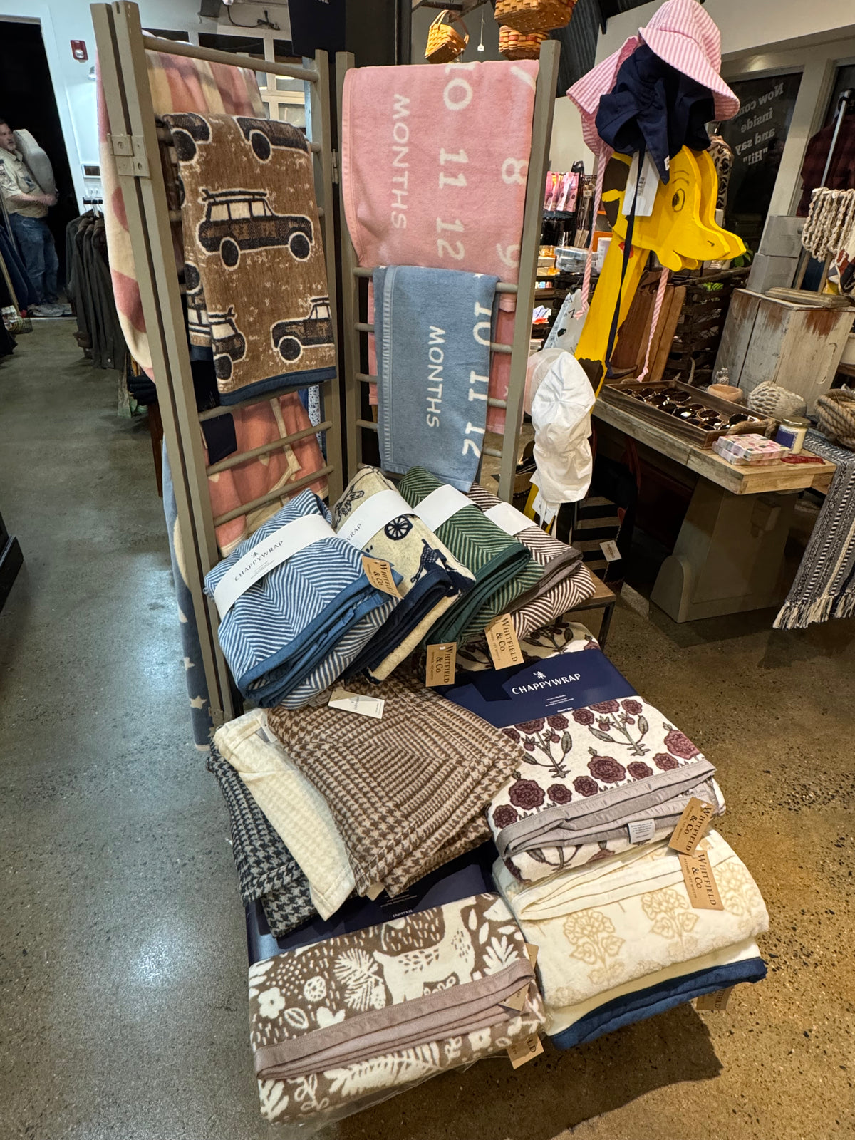 A retail display rack filled with folded and hanging baby blankets and swaddle blankets in various patterns and colors, including brown, pink, blue, green, and cream. The brand name 'Eagles Nest' is visible on several navy blue tags.