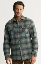 A man wearing a long-sleeve button-up plaid shirt in green, black, and white, paired with brown pants, photographed against a light gray studio background.
