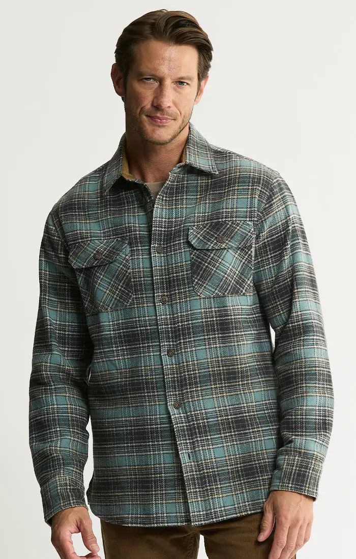A man wearing a long-sleeve button-up plaid shirt in green, black, and white, paired with brown pants, photographed against a light gray studio background.