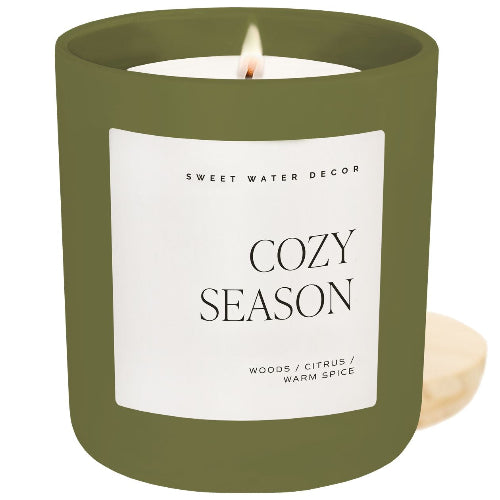 A lit candle in an olive green glass jar with a white label reading 'COZY SEASON' and 'SWEET WATER DECOR'. A small beige coaster is visible on the right against a white background.