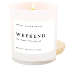 A lit white glass candle jar with a white label reading 'SWEET WATER DECOR WEEKEND' and a small wooden lid resting behind it on a white background.
