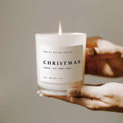 Hand holding a lit Sweet Water Decor Christmas scented soy candle in a glass jar, with a neutral background.