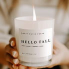 A lit white scented candle in a frosted glass jar held in hands. The front label reads 'SWEET WATER DECOR' and 'HELLO FALL' with 'CIDER + RUMEG + CINNAMON + CLOVES' below, against a softly blurred warm neutral background.