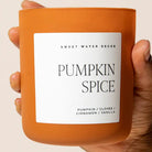 A hand holding a matte orange candle jar labeled 'PUMPKIN SPICE' by Sweet Water Decor, with a white label and black text on a light beige background.