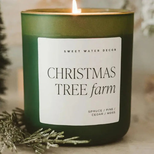 A lit green candle in a matte green jar with a white label reading 'SWEET WATER DECOR CHRISTMAS TREE farm' and 'SPRUCE / PINE / CEDAR / MOSS' near the bottom. The candle is styled with frosted pine branches on a soft neutral background.