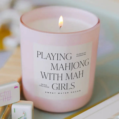 A pink glass candle with a lit flame and a white label reading 'PLAYING MAHJONG WITH MAH GIRLS' and 'SWEET WATER DECOR,' styled with mahjong tiles in the foreground.