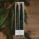 A boxed set of two tree-shaped taper candles displayed on a rustic wooden surface with pine branches, pinecones, and evergreen foliage around it. The package label reads 'SWEET WATER BAKERS' and 'TREE taper candles'.