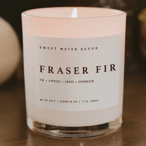 A lit glass jar candle labeled 'SWEET WATER DECOR FRASER FIR' on a dark wooden surface, with a softly blurred warm-toned background.