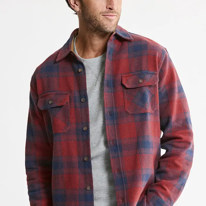 A man wearing a red and navy blue plaid flannel shirt over a gray crewneck t-shirt, photographed against a clean white background.