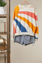 A long-sleeve crewneck sweater with diagonal stripes in yellow, coral pink, blue, and white, styled with distressed denim shorts on a wooden hanger against a neutral background.